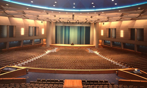 Thousands of wood-baked auditorium seats face a stage covered by a blue curtain in an enormous performance hall. The ceiling is painted with a blue star-burst pattern.
