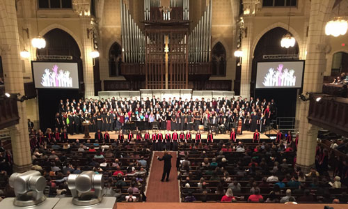 A large choir stands before a church full of people. A silver and gold church organ rises behind them with two projection screens on either side.