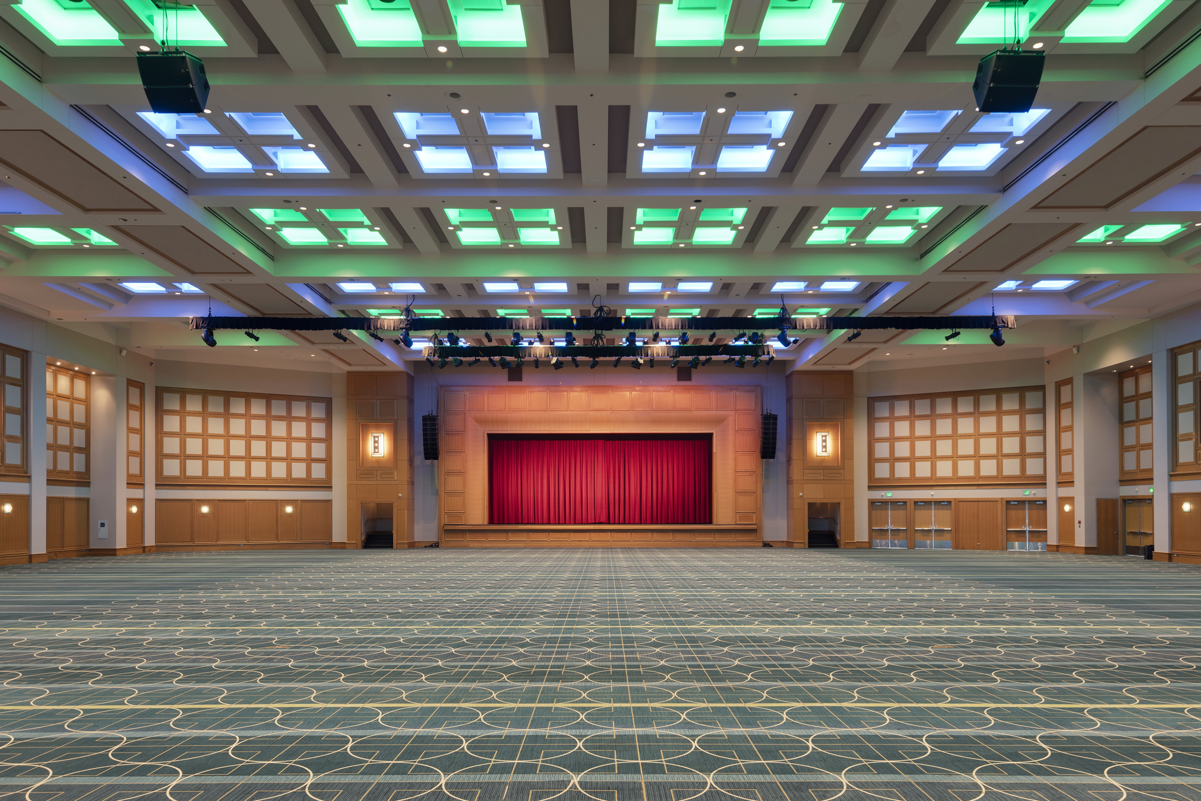 A large, empty ballroom with patterned carpet and blue and green lights on the ceiling leads to a wood-paneled stage covered with a red curtain.