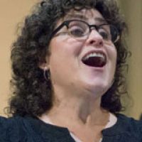 headshot of a white woman with black curly hair and glasses and her mouth wide open