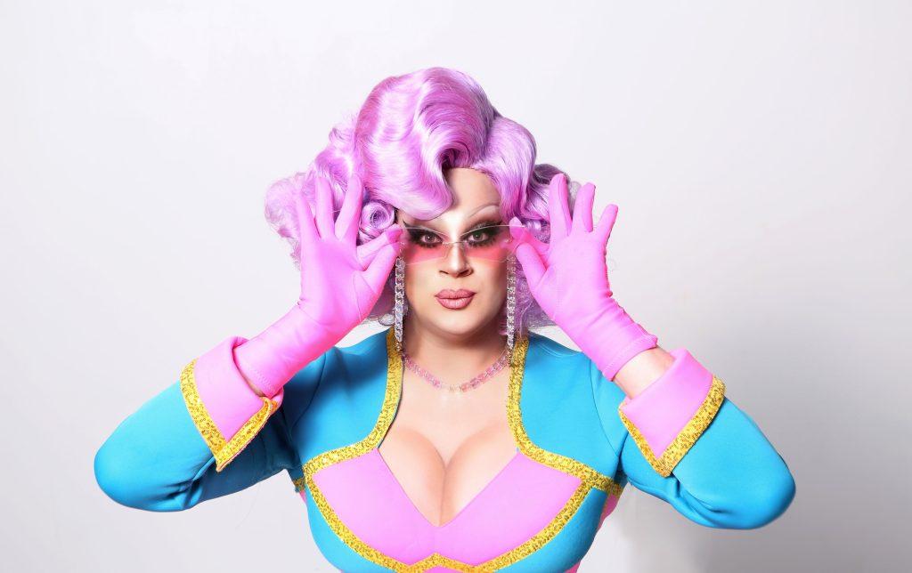 Nina West in gorgeous blue and purple drag with gold lining, wearing a wavy purple wig