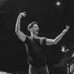 A black and white candid of Brayton conducting with a dynamic pose, in a black t-shirt and jeans