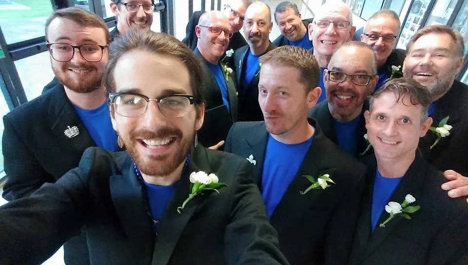 A group of singers with short hair, blue shirts, and dark blazers with pinned white flowers