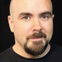 A headshot of a bald male-presenting person with a short beard and mustache - smiling slightly.