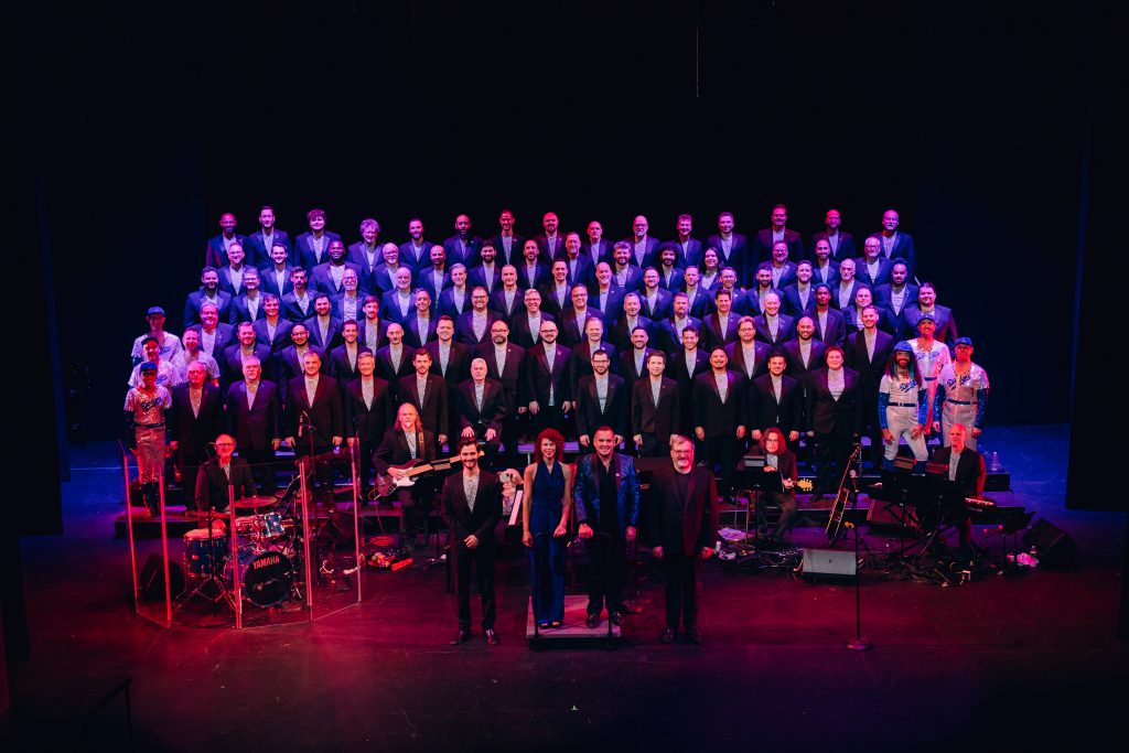 A chorus on risers with male presenting singers bathed in purple light