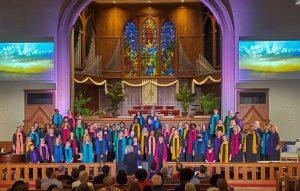 The Atlanta Women's Chorus stands at the front of a Church, wearing black and multicolored scarves.