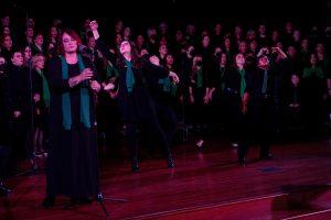 The Denver Women's Chorus stands on a stage behind two dancers and a soloist at a microphone. Everyone wears black with green scarves.