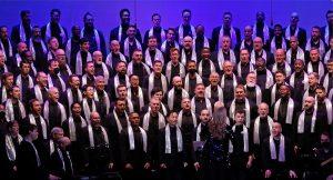 The Gay Men's Chorus of Washington, DC, sings wearing black with silver scarves in front of a purple background. Artistic Director Thea Kano stands with her back to us, facing the choir.