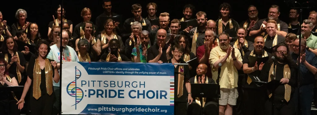 The members of Pittsburgh Pride Choir stand on risers and applaud behind a banner with the chorus's logo.
