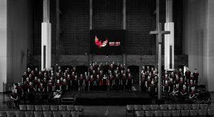 The Twin Cities Gay Men's Chorus stands on risers in a church. The only color outside of black and white is the red writing on their shirts and a projection reading 