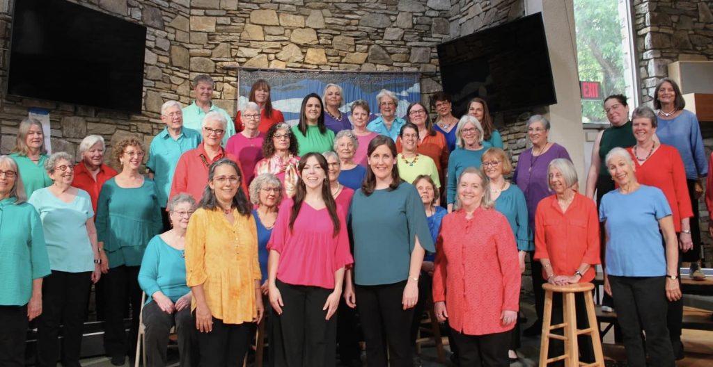 The members of Womansong stand before a brick wall wearing multicolored shirts and black pants