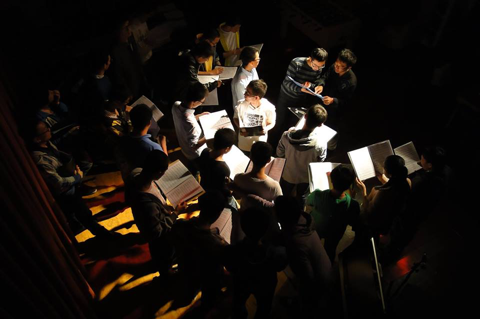 Singers in a dark room hold binders as notes are given.