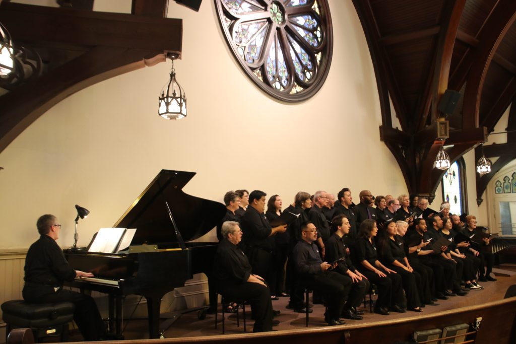 A choir performs on a stage with an accompanist, some members sit while others stand.