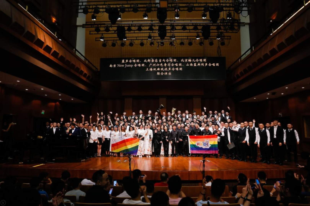 A group of people wearing formal wear and holding pride flags stand on a stage posing for a picture