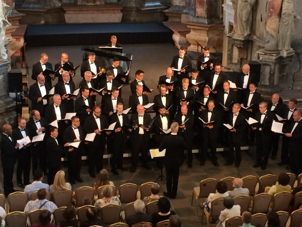 A men's chorus in suits perform on a stage before an audience