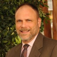 Eric smiles through a salt and pepper beard, wearing a brown suit and red tie.