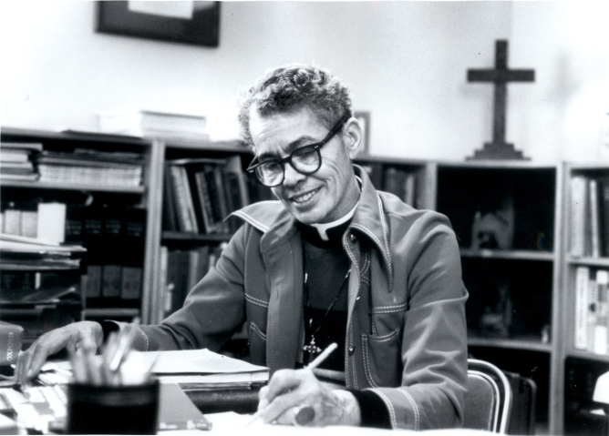 In black and white, Pauli Murray sits at a desk writing, wearing clerics with a cross on the wall behind.
