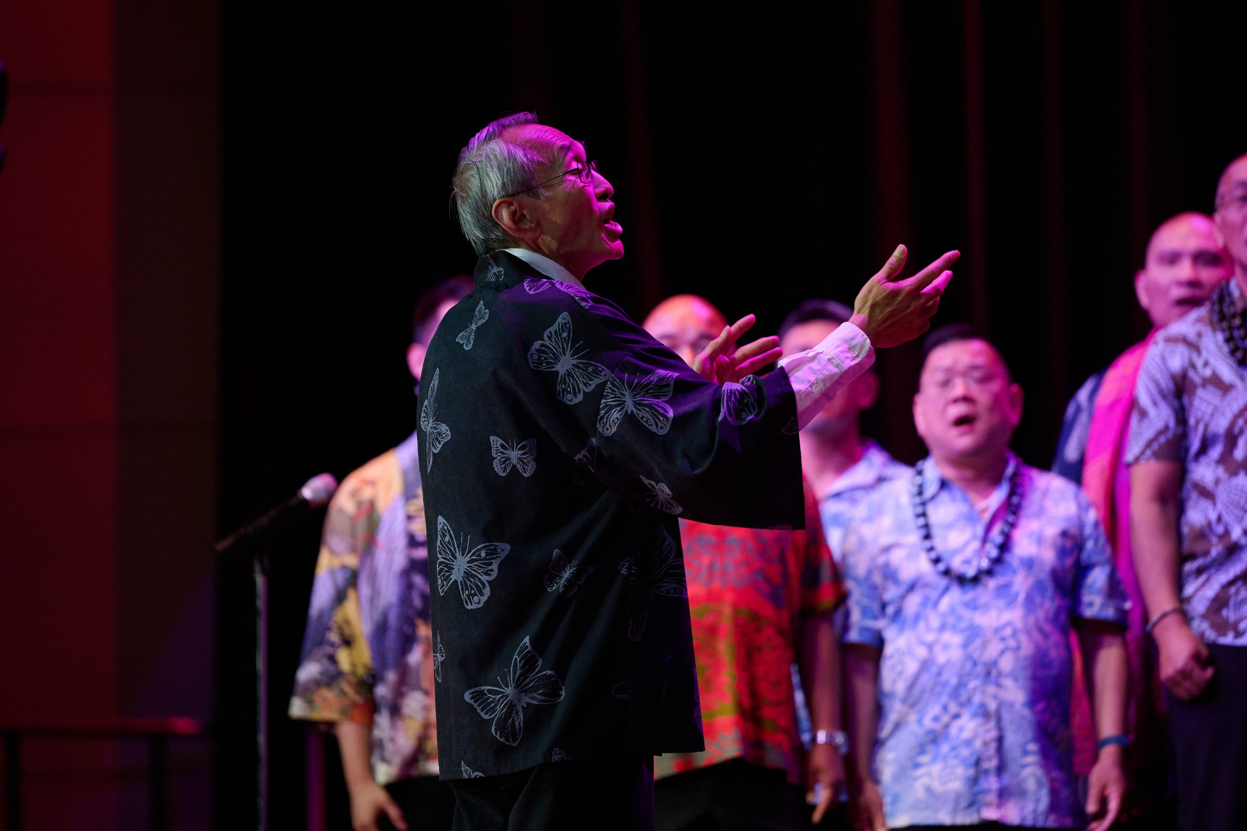 Randall Kikukawa conducts GAPA Men's Chorus at Festival 2024 in Minneapolis, he is bathed in purple light.