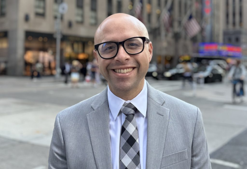 John Carrion stands on a street corner in NYC smiling. He wears a grey suit and dark rimmed glasses.