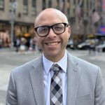 John Carrion stands on a street corner in NYC smiling. He wears a grey suit and dark rimmed glasses.