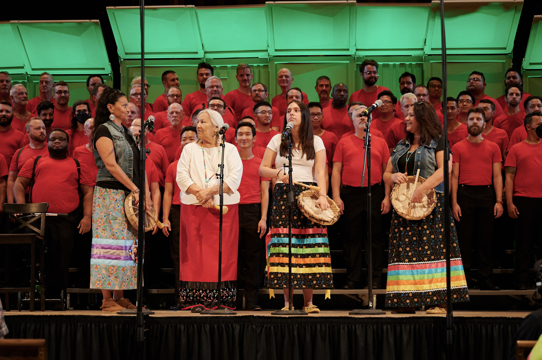 Four guest artists with hand-held drums stand in front of a chorus wearing red shirts at Festival 2024.