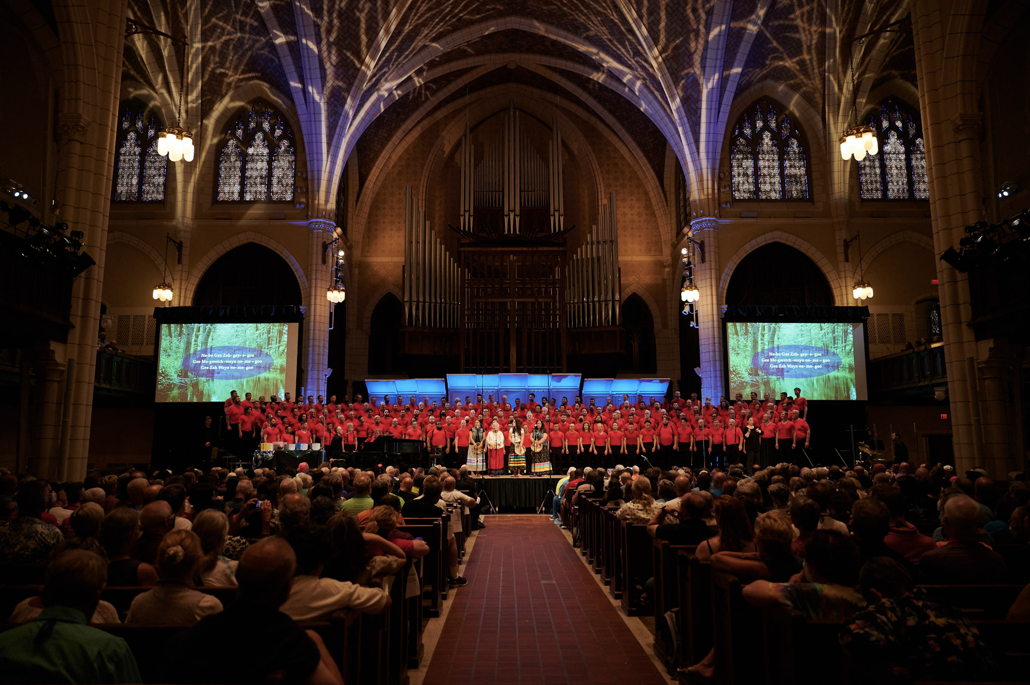 Four guest artists with hand-held drums stand in front of a chorus wearing red shirts at Festival 2024 at Central Lutheran Church.