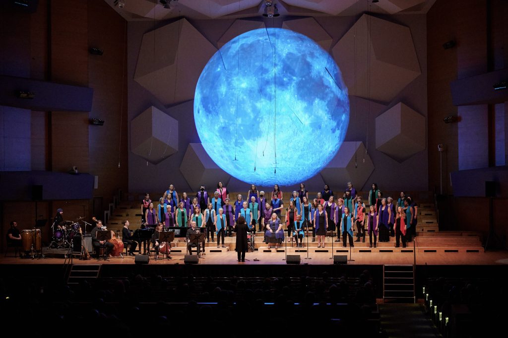 Anna Crusis Feminist Chorus performs in Orchestra Hall at Festival 2024, a large blue moon glows on the wall behind them.