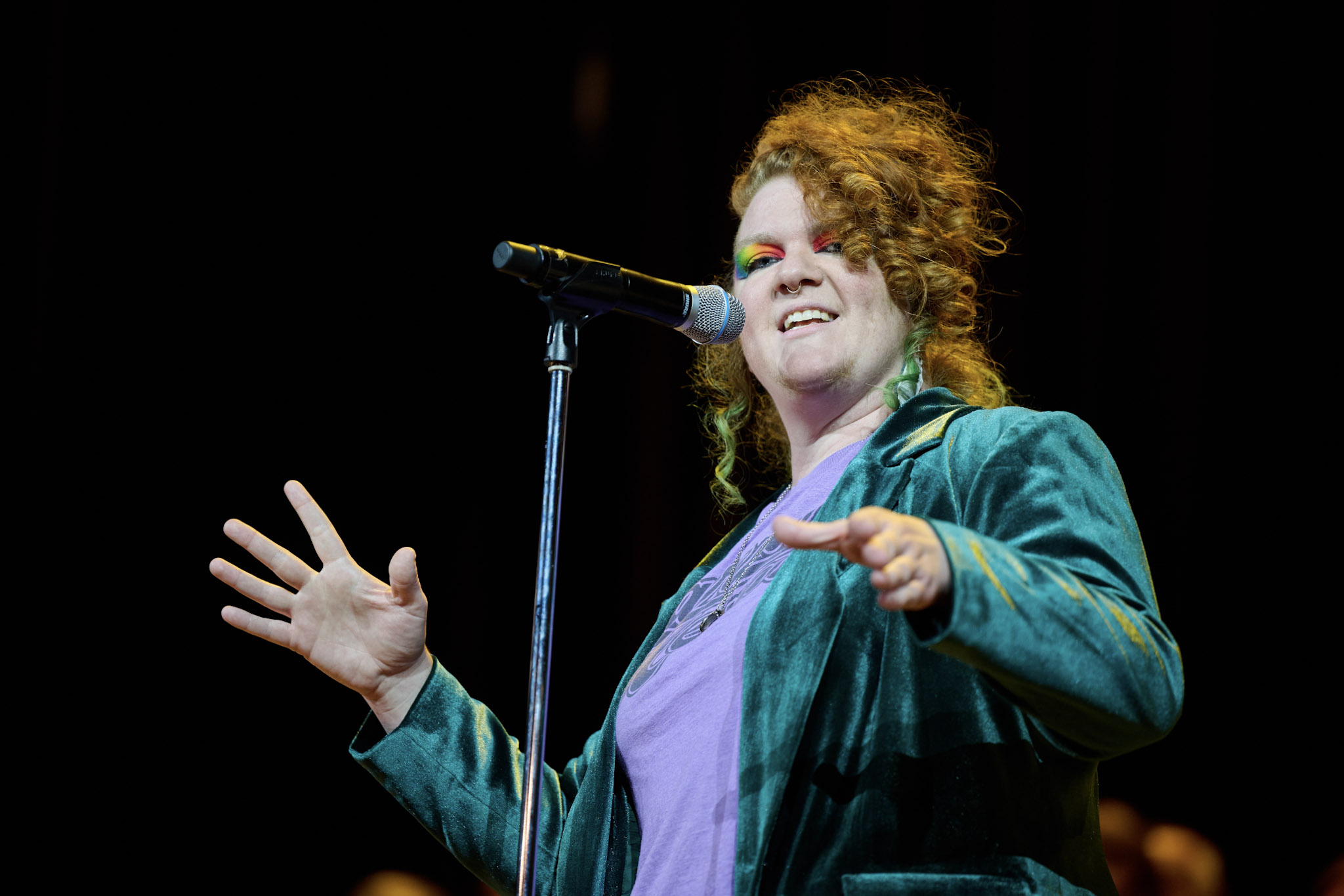 A performer wearing a velvet blazer with long, curly red hair and rainbow eye shadow sings into a microphone at Festival 2024.