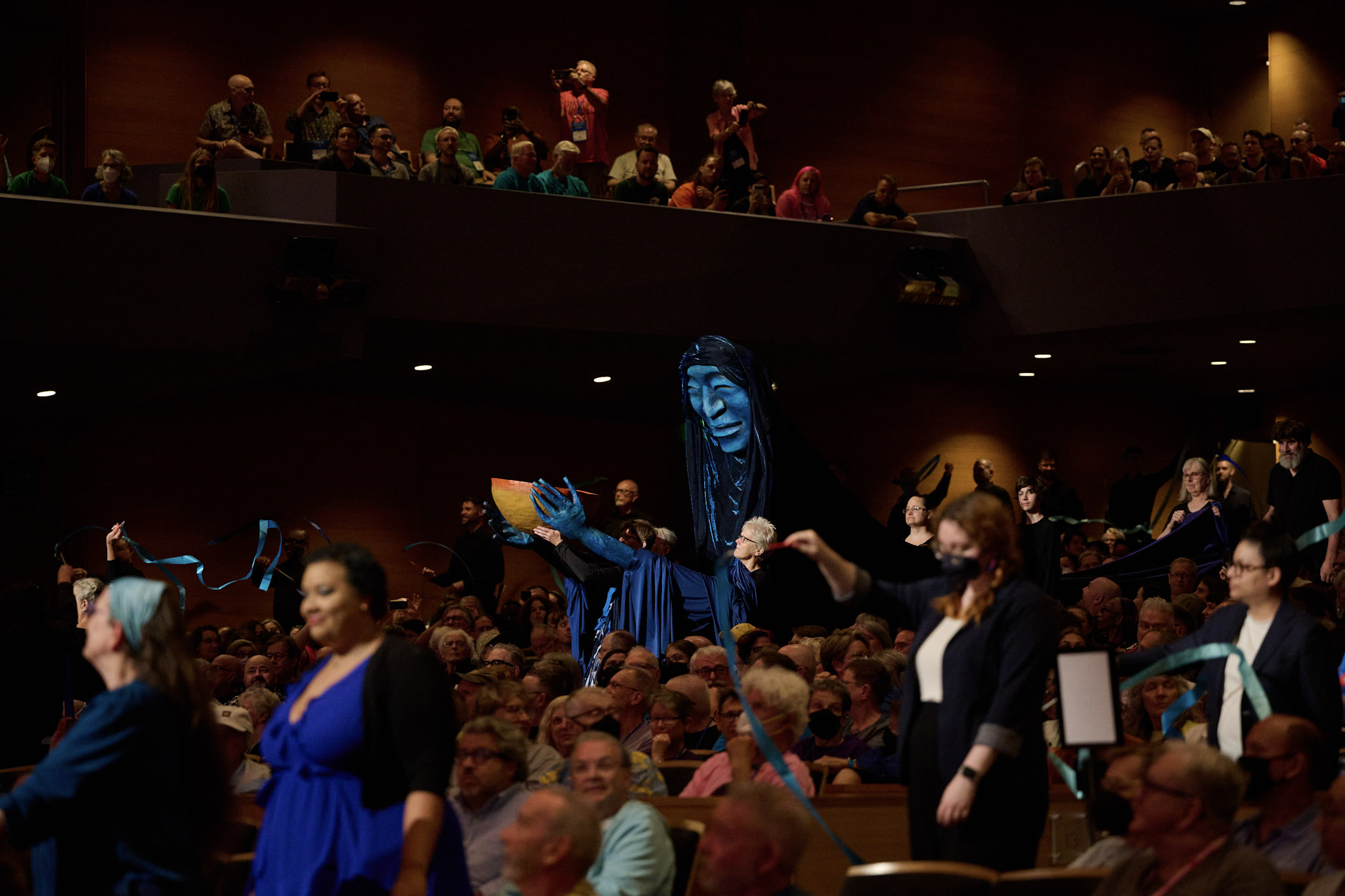 A large blue puppet moves down the aisle of a crowded Orchestra Hall at Festival 2024 in Minneapolis.