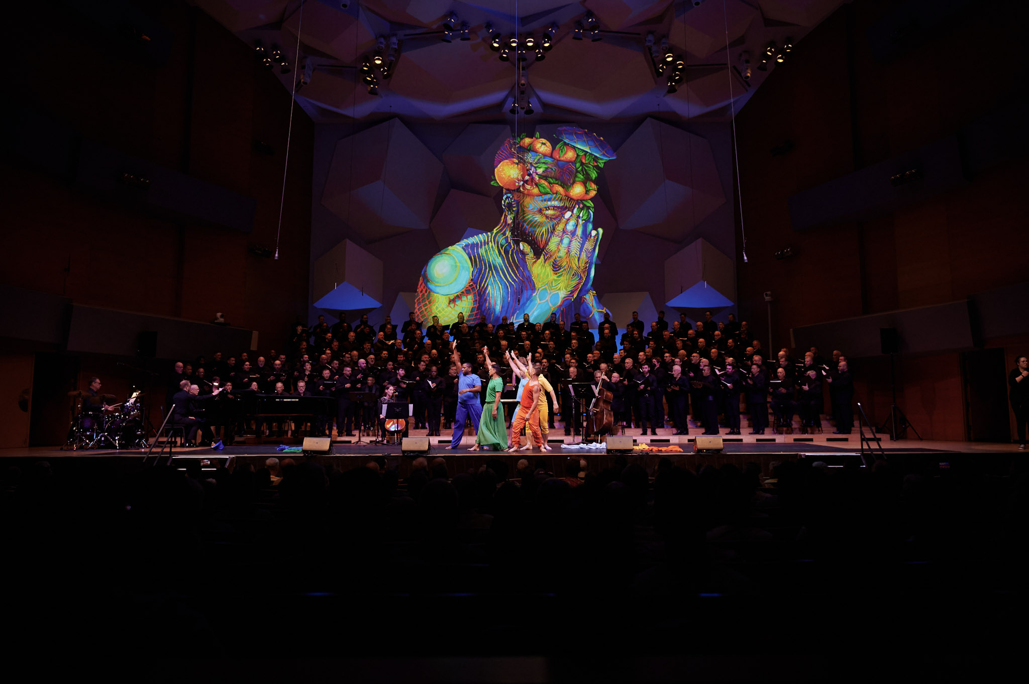 The Gay Men's Chorus of Washington, DC, performs in Orchestra Hall at Festival 2024. A colorful portrait is projected on the back wall with a group of colorfully-dressed dancers downstage.