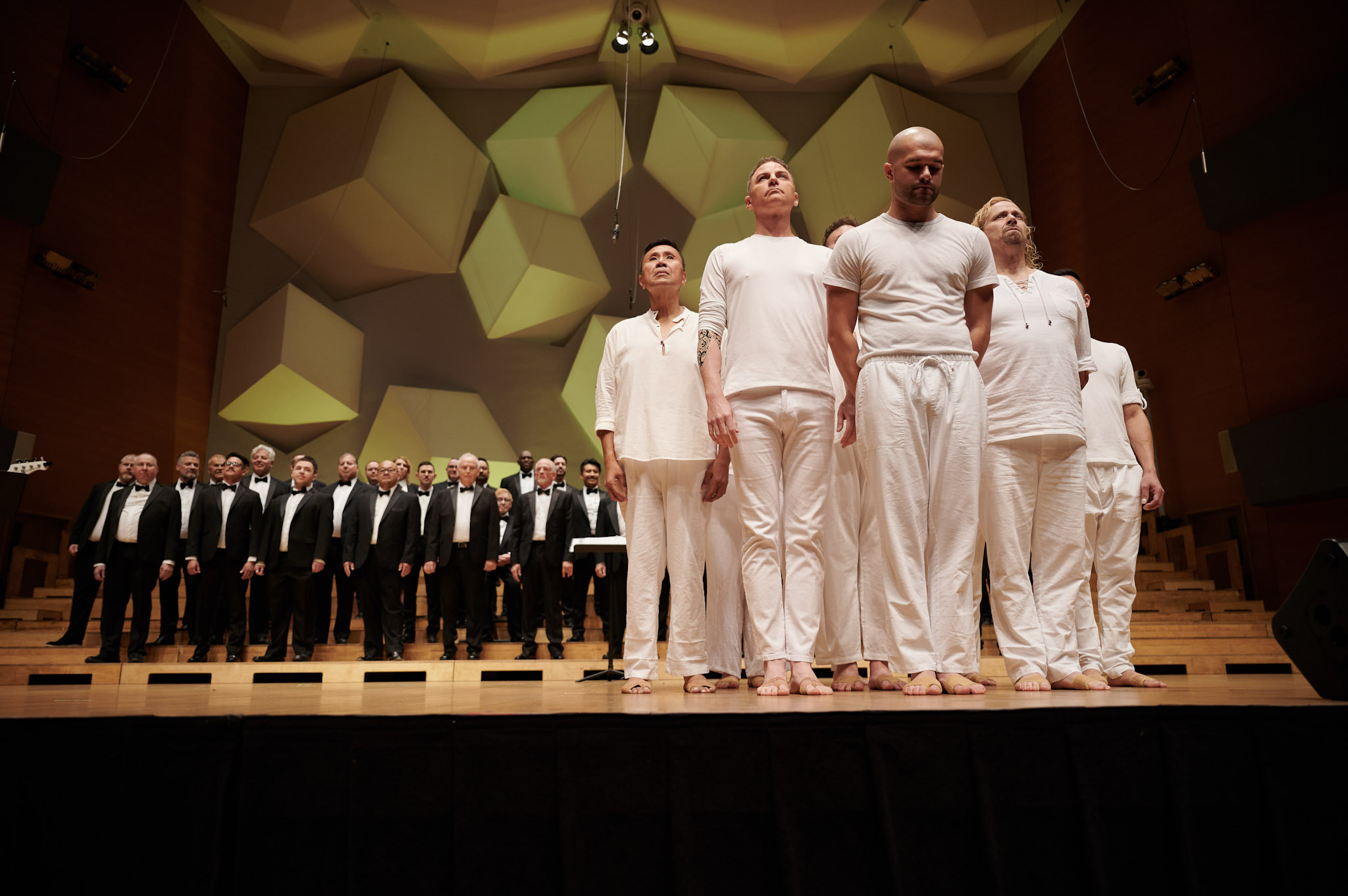 A choir performs at Orchestra Hall wearing black tuxes in front of a group of dancers dressed in white at Festival 2024.