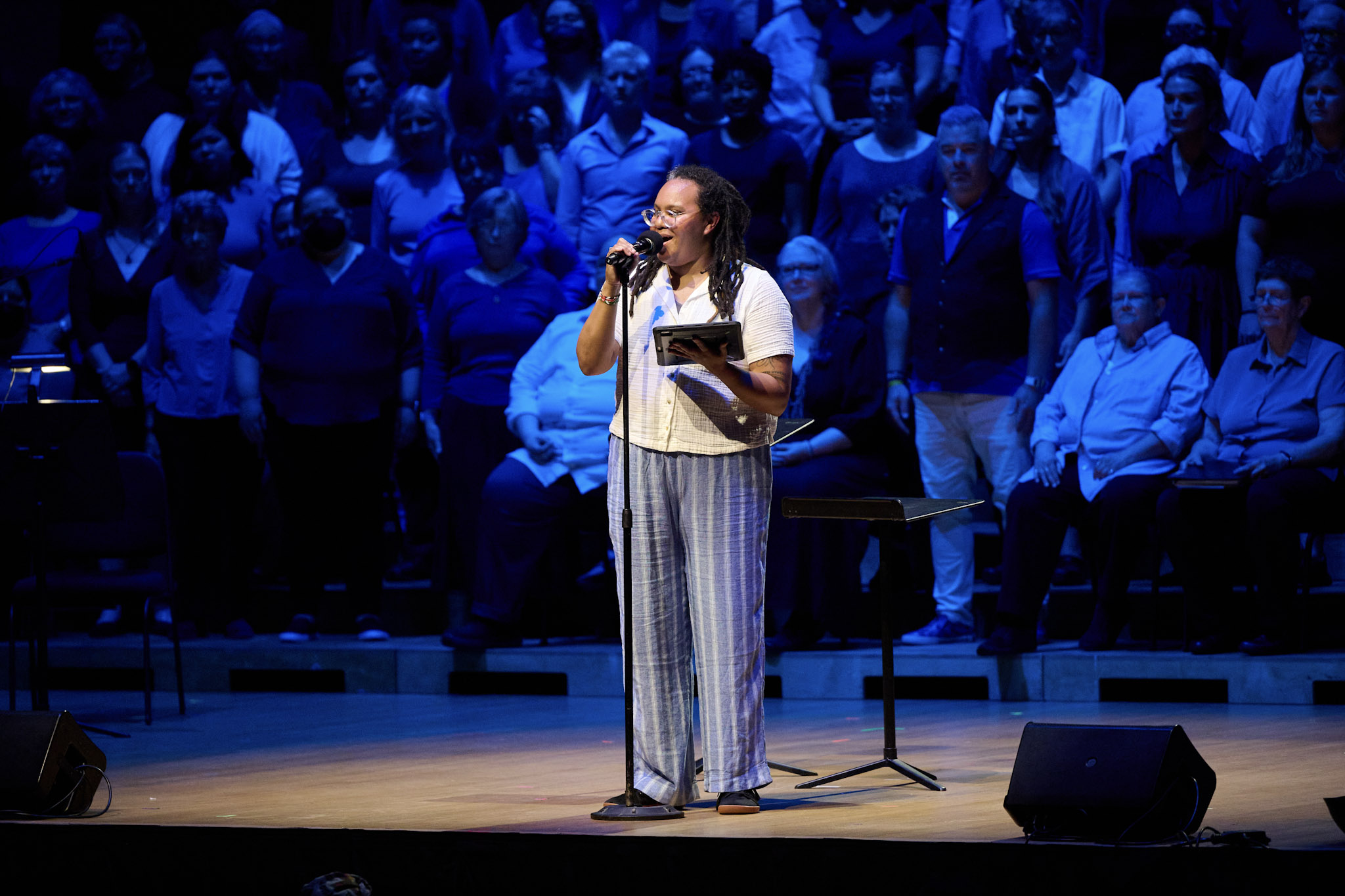 Waigwa speaks into a microphone in front of One Voice Mixed Chorus lit in blue.