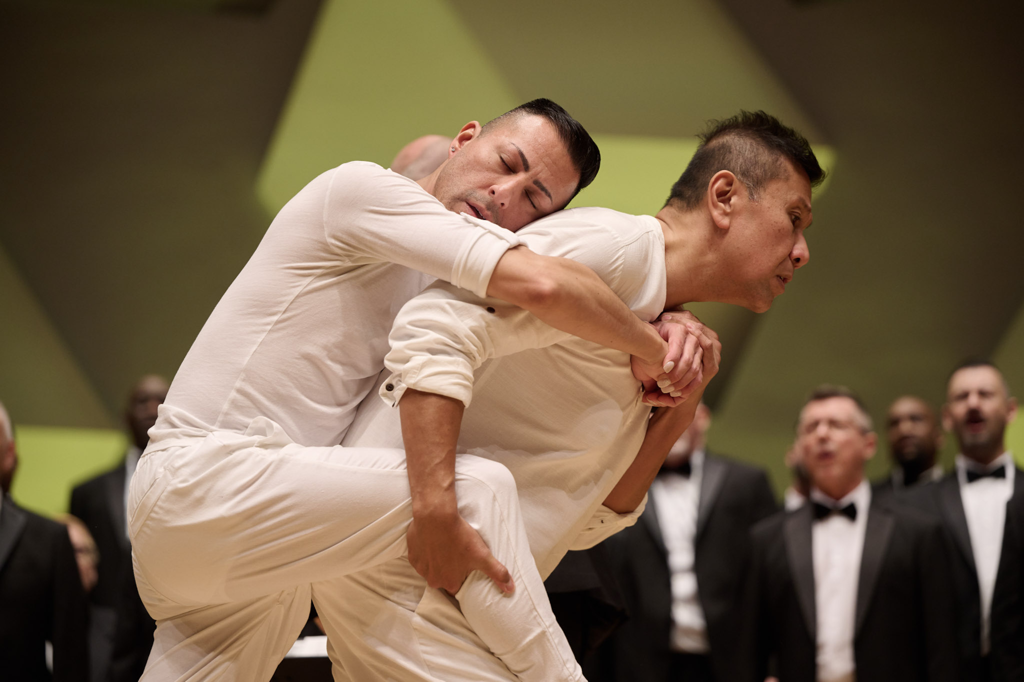 A choir performs at Orchestra Hall wearing black tuxes in front of two dancers dressed in white at Festival 2024.