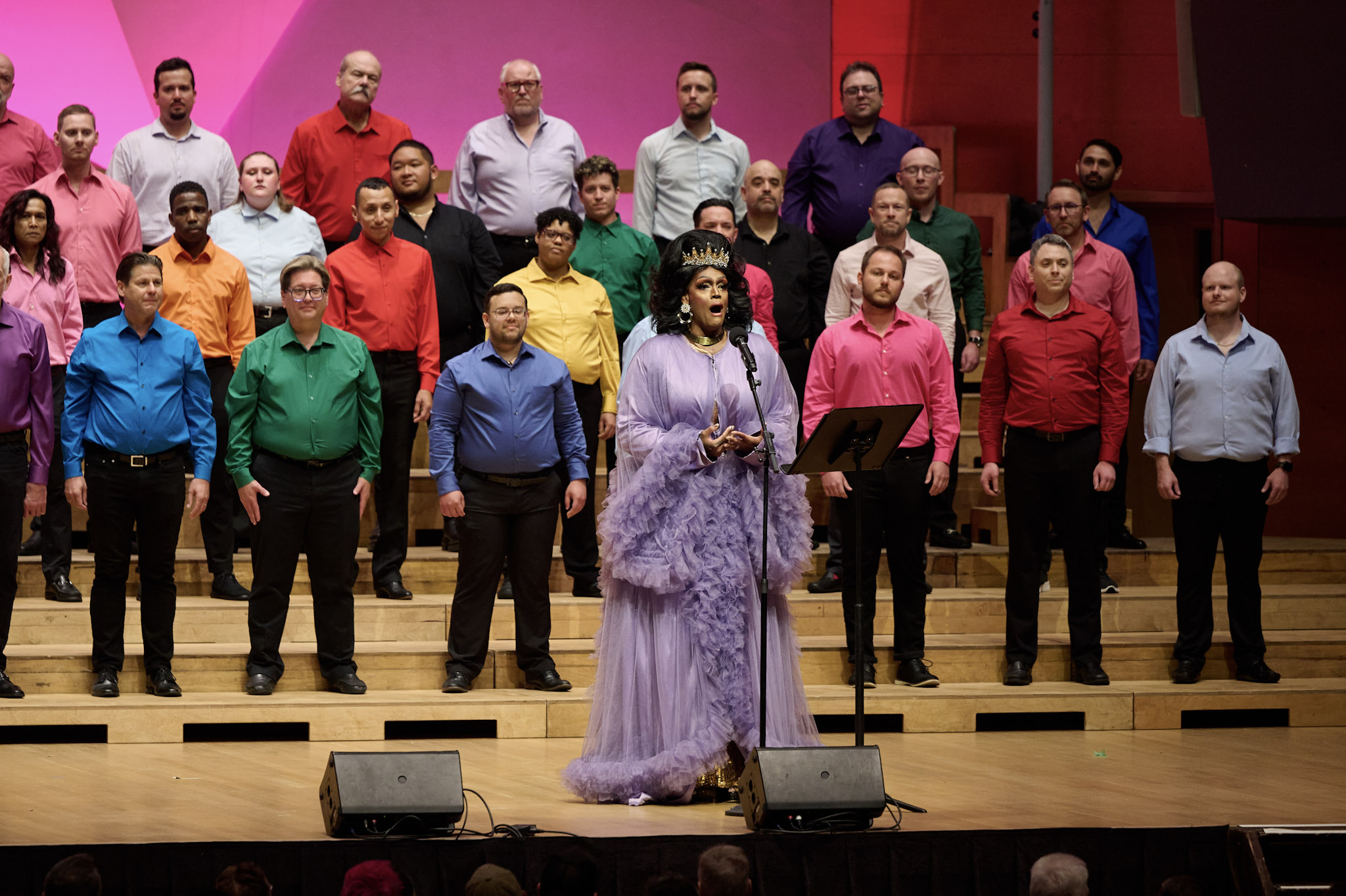 A drag queen dressed a filly purple gown sings in front of a colorfully dressed choir at Festival 2024.