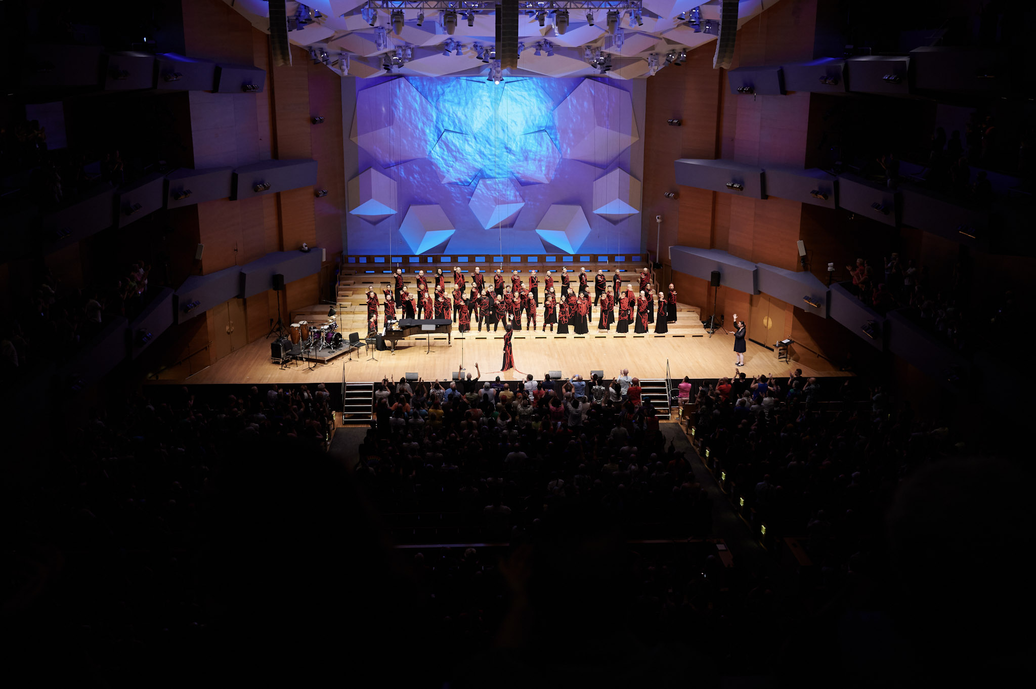 Coro Ciudad de Mexico performs at Festival 2024, wearing black with red rope and yarn covering and connecting them, water is projected on the wall behind them.