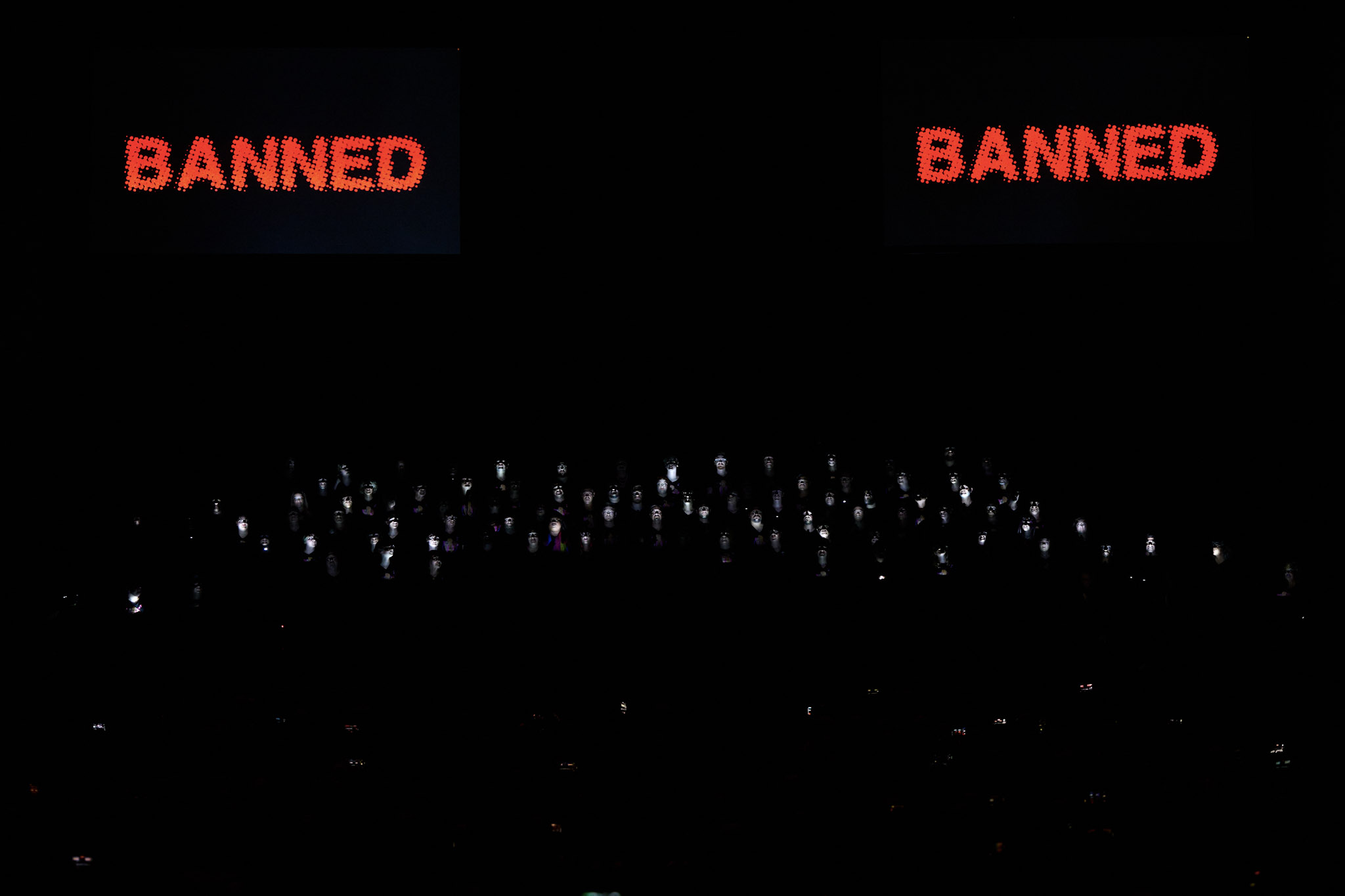 A choir performs in the dark with just their faces illuminated at Festival 2024 the word "banned" appears twice in red letters above them.