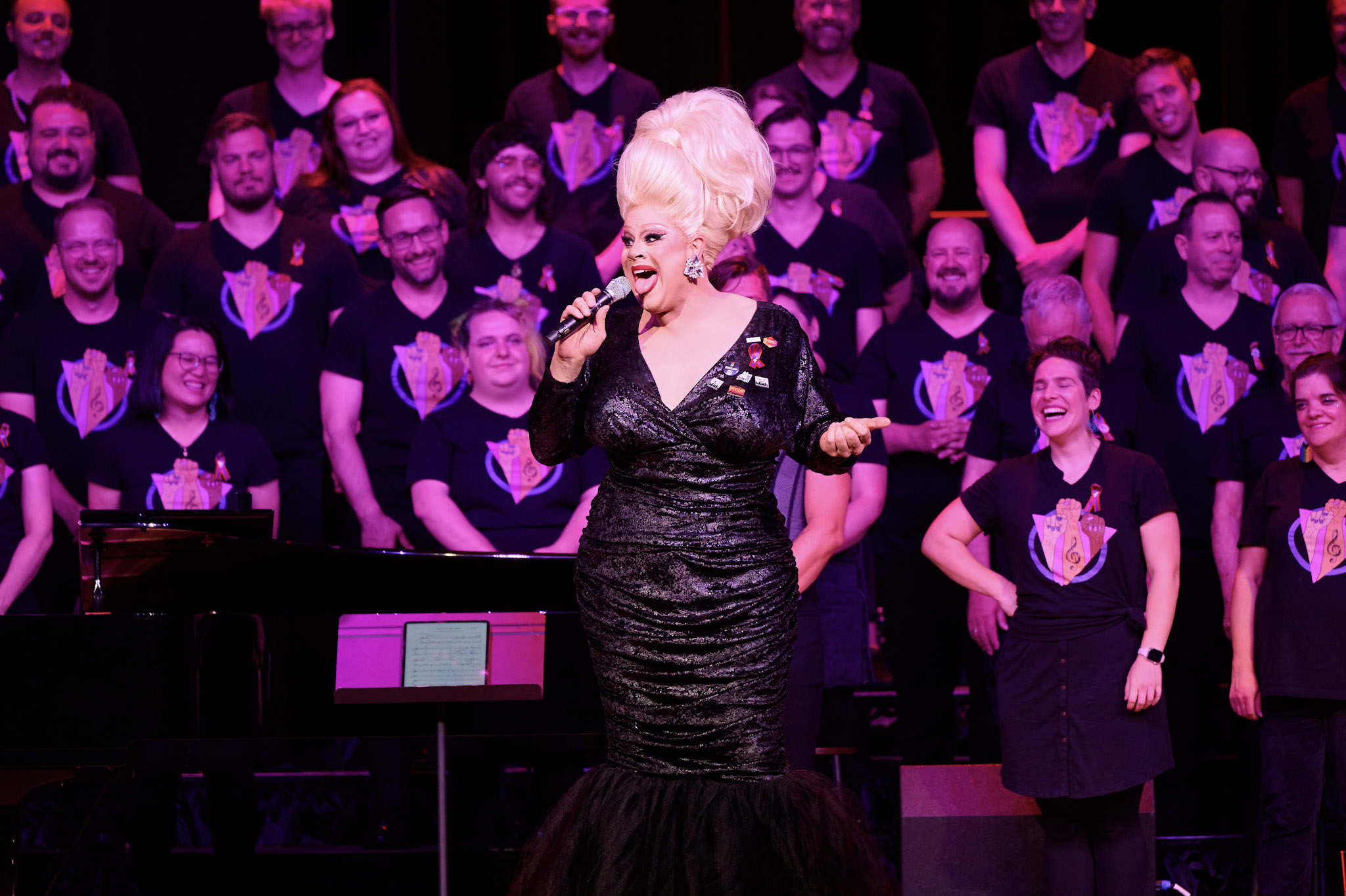 A drag queen with a tall white beehive and black dress performs at Festival 2024 in front of choir.