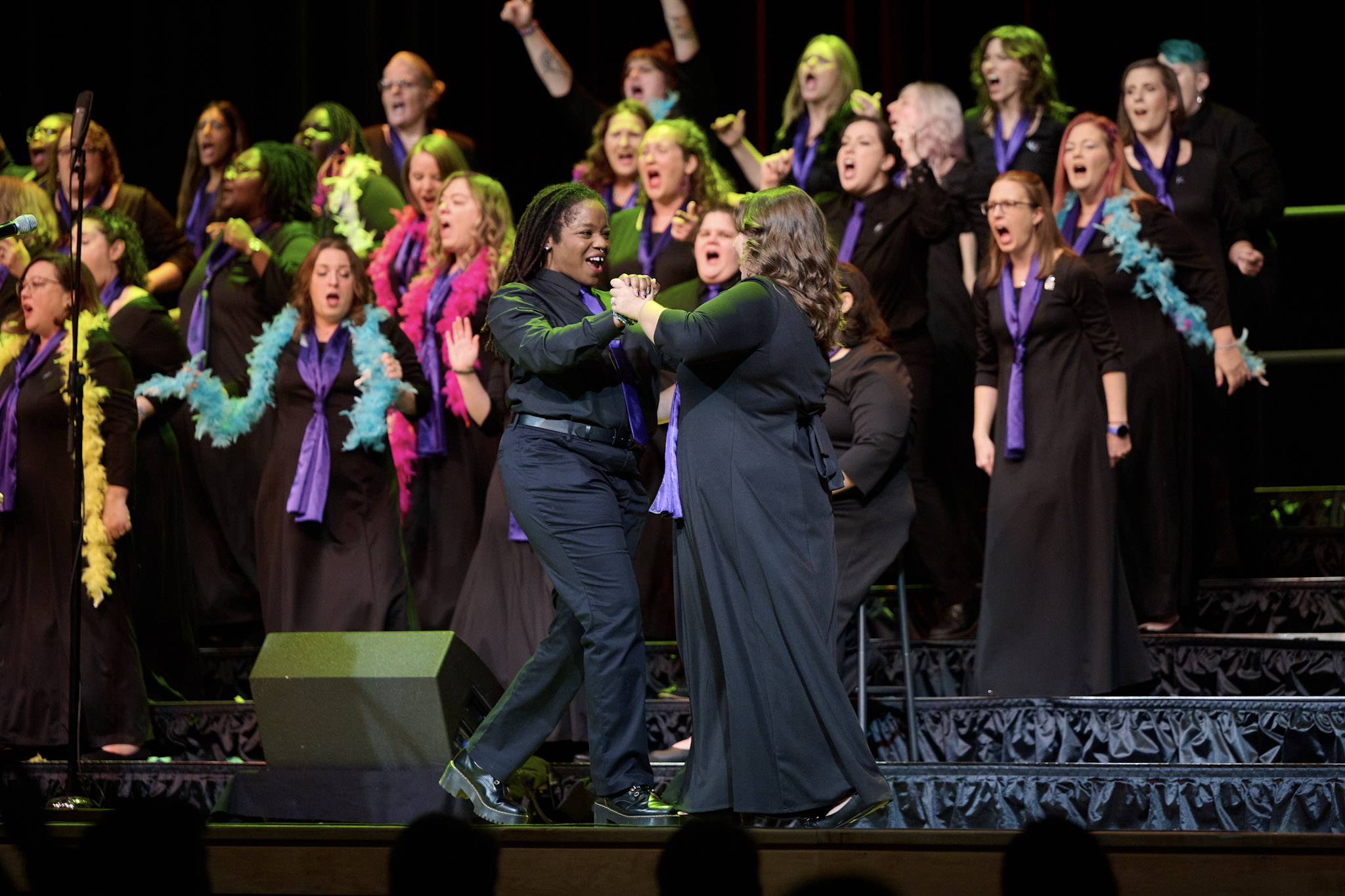 Two singers wearing black dance in front of a choir wearing colorful boas at Festival 2024.