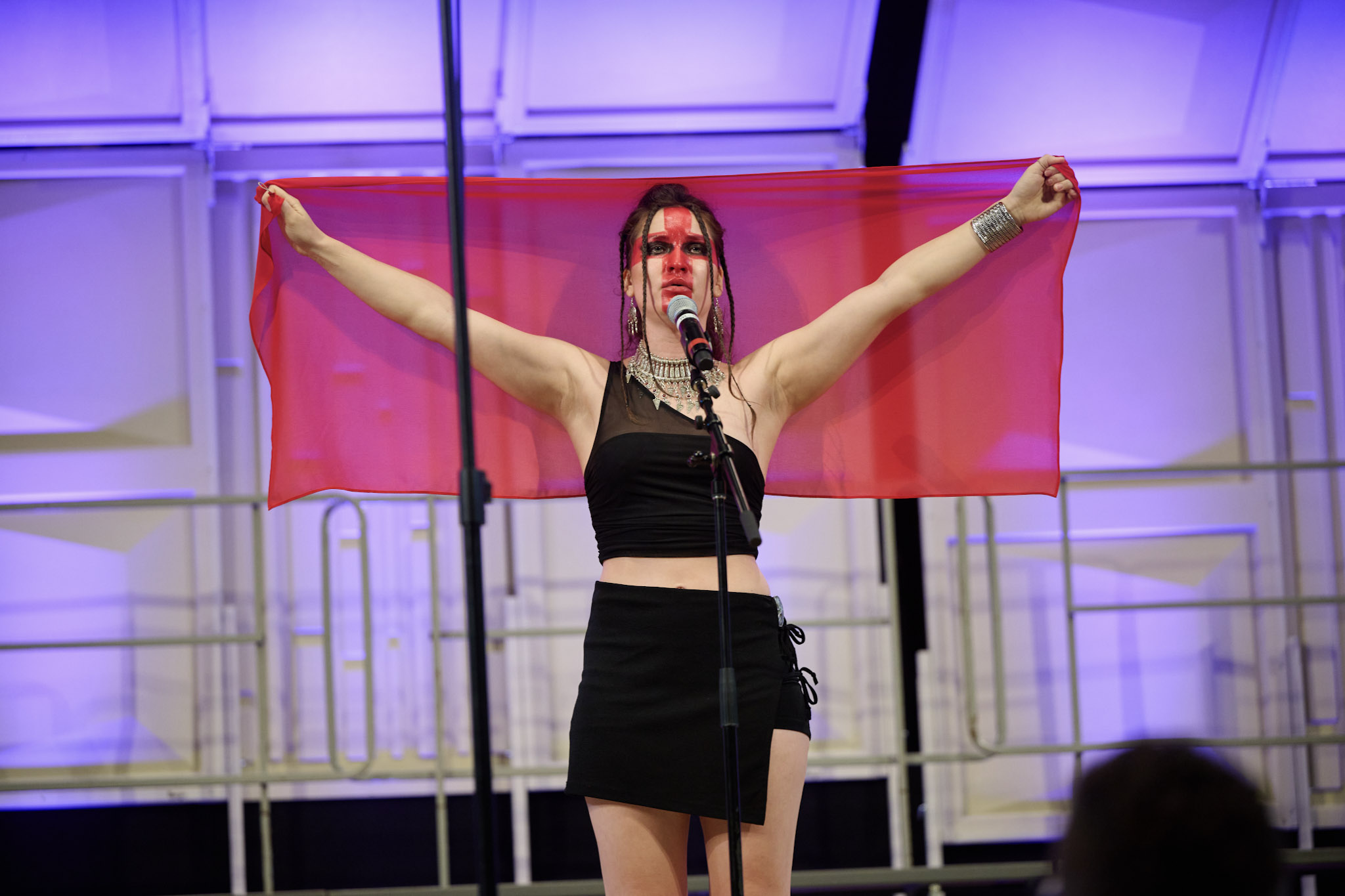 A performer holds up a red translucent cloth behind their head with a red cross painted across their face at Festival 2024.