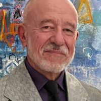 Cliff Schiappa, a white man with silver facial hair wears a suit in front of a colorful background.