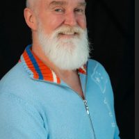 Will Jones, a white man with silver hair and a long white beard, wears a blue shirt in front of a black background.