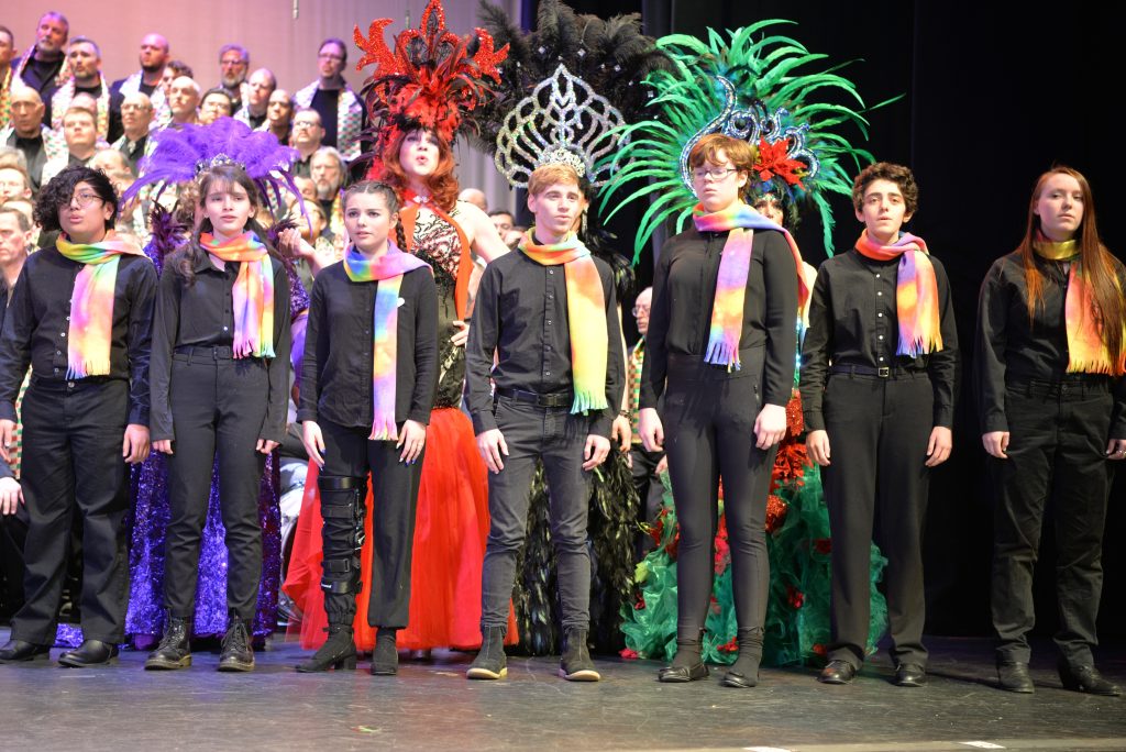 Members of GenOUT Youth Chorus sing with the Gay Men's Chorus of Washington, DC, they wear black with colorful scarves in front of feathered drag queens.