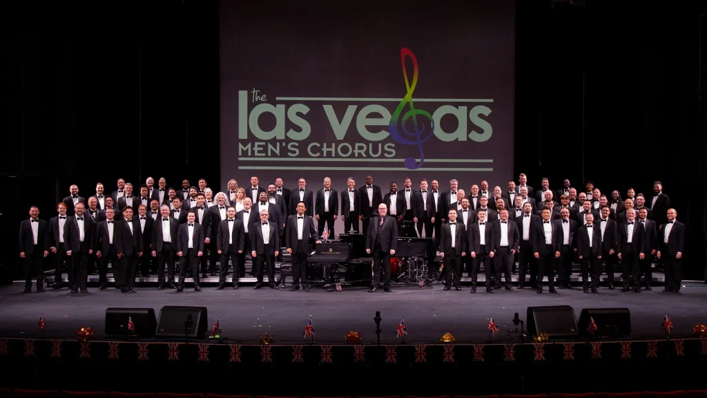 Las Vegas Men's Chorus stands on stage wearing tuxedos below their projected logo.