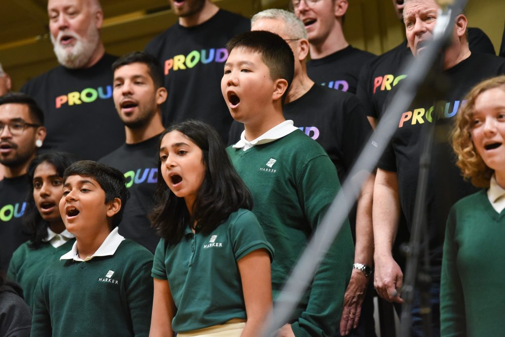 Students wearing green sweaters sing with members of SFGMC wearing black shirts with the word "PROUD" written in rainbow letters.