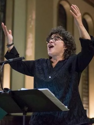 Miriam Davidson raises her arms while directing and singing. She is a white woman with dark curly hair, wearing a black button-up shirt.