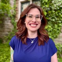 Portrait of Dr. Jessica L. Gallagher-Steuver, a white woman with shoulder-length auburn hair, clear-rimmed glasses, and a blue shirt outside of an ivory-covered brick wall.